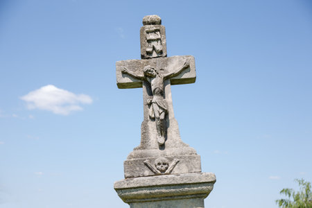 Slightly inclined ancient stone tombstone in the form of the cross with crucified Jesus sculpture on the old Christian cemetery against the sky in sunny dayの写真素材