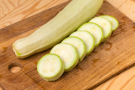 Sliced into circles light green vegetable marrow against the same whole marrow on the wooden cutting board on the rustic table, close-up in selective focusの写真素材
