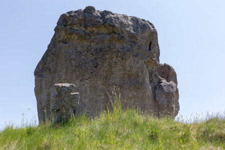 Old stone cross among the grass on the medieval Cossack grave on the hill against the big single sandstone rock in sunny dayの写真素材
