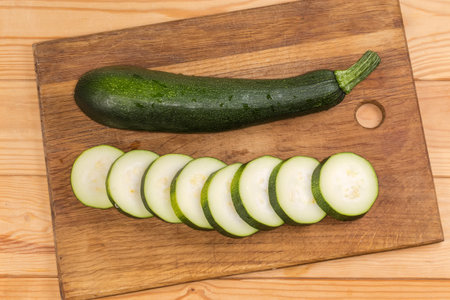 One dark green whole zucchini and sliced into circles the same zucchini on the wooden cutting board on the rustic table, top viewの写真素材