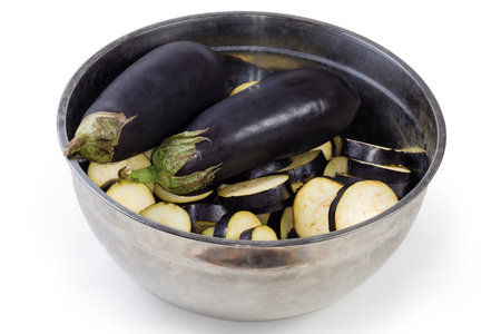 Ripe raw whole and sliced into circles purple eggplants in big stainless steel kitchen bowl on a white backgroundの写真素材