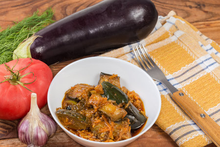 Eggplant pieces stewed with other vegetables in the bowl against the raw ingredients for their preparation on the rustic tableの写真素材