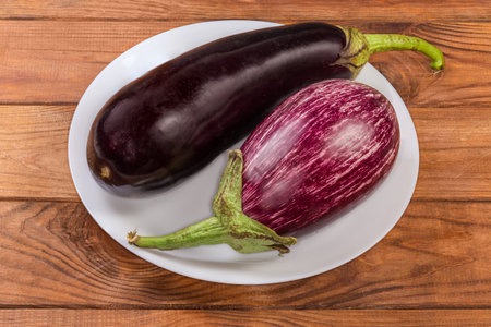 One raw whole conventional eggplant and one striped, so-called Graffiti eggplant on the white dish on a rustic tableの写真素材