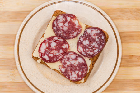 Open sandwich with slices of the dry-cured sausage and cheese on the saucer on the cutting board, top viewの写真素材