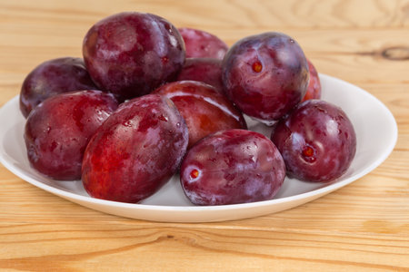 Fresh ripe washed plums covered with water drops on the white dish on the rustic table, side view close-up in selective focusの写真素材