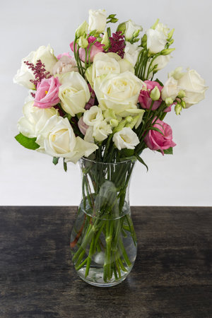 Bouquet of white and pink roses and white eustoma in glass transparent flower vase with water standing on black surface on a light backgroundの写真素材