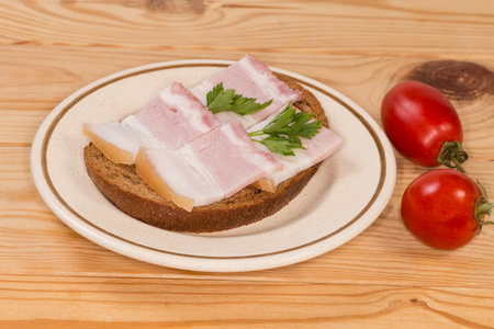 Open sandwich with slices of the boiled-smoked pork belly on skin decorated with fresh parsley twigs on saucer and cherry tomatoes on the rustic table, side view close-upの写真素材