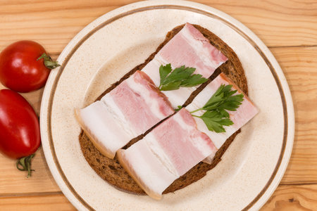 Open sandwich with slices of the boiled-smoked pork belly on skin decorated with fresh parsley twigs on saucer and cherry tomatoes on the rustic table, top view close-upの写真素材