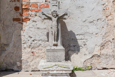Ancient stone sculpture of the crucified Jesus next the mediaeval defensive stone and bricks wall with partly collapsed plaster in sunny dayの写真素材