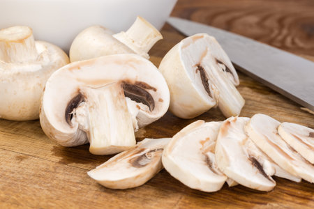 Whole, halved and sliced button mushrooms on the cutting board against the kitchen knife, view close-up in selective focusの写真素材
