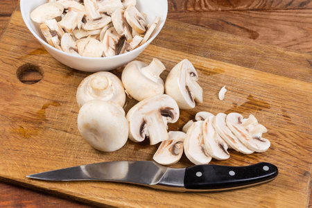 Sliced button mushrooms in the small bowl, partly sliced the same mushrooms and kitchen knife on the cutting board on the old wooden rustic tableの写真素材