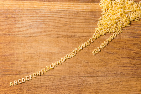 Raw pasta in the shape of capital letters of English alphabet and Arabic numerals placed in line in the alphabetical order stretched out from a heap on the cutting boardの写真素材