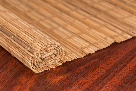 Partially rolled place mat woven from thin natural wooden bamboo strips and cotton string lies on the red wooden table, fragment close-up in selective focusの写真素材