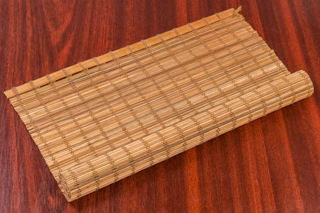 Partially rolled place mat woven from thin natural wooden bamboo strips and cotton string lies on the red wooden tableの写真素材