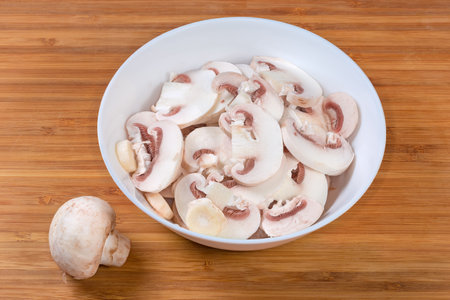 Fresh raw button mushrooms chopped into thin slices in white bowl on a wooden surfaceの写真素材