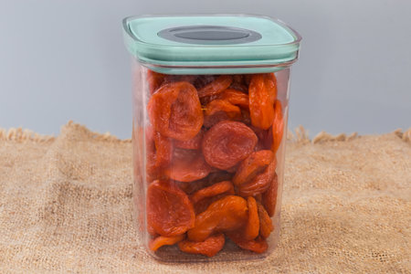Dried smoked apricots in transparent container with closed lid on a surface covered with a burlap, side view on a gray backgroundの写真素材