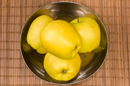 Whole big yellow apples in the stainless steel kitchen bowl on a bamboo table mat, top viewの写真素材