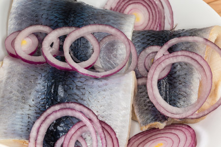Pickled herring fillets on skin with fresh red onion sliced into rings on the white dish, top view close-upの写真素材