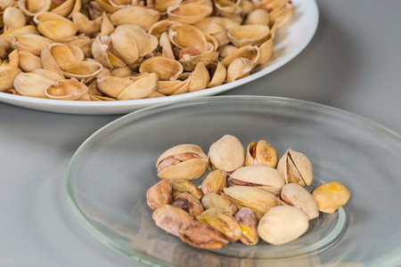 Partly peeling roasted salted pistachio nuts on the glass saucer against the empty shells on a big dish on a gray background, fragment side view in selective focusの写真素材