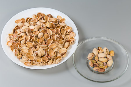 Partly peeling roasted salted pistachio nuts on the glass saucer against the empty shells on a big dish on a gray backgroundの写真素材