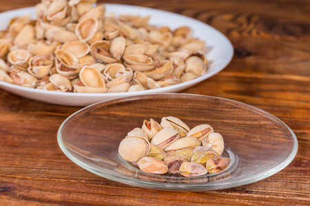 Partly peeled roasted salted pistachio nuts on the glass saucer on a blurred background of the empty shells on a big dish on a rustic table, side view in selective focusの写真素材