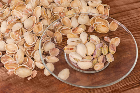 Partly peeled roasted salted pistachio nuts on the transparent glass saucer against the empty shells on a rustic tableの写真素材