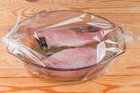 Two raw gutted redfish without heads, or ocean perch prepared for baking in the old glass baking dish placed in the plastic heatproof oven bag on a rustic tableの写真素材