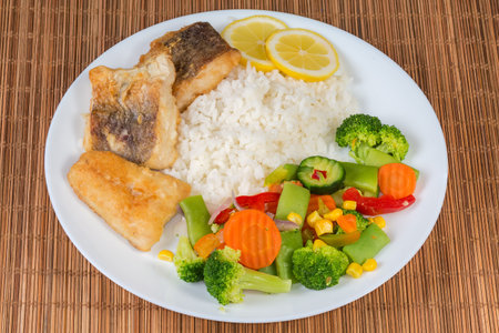Fried hake fillets pieces with garnish of the boiled vegetables mix and rice on a white dish on a bamboo place matの写真素材