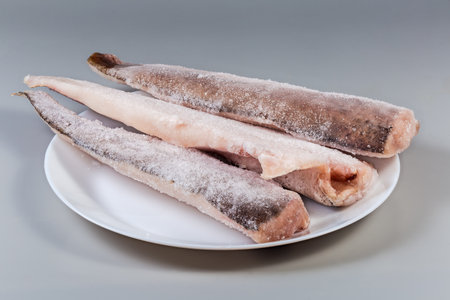 Raw frozen fillets of hake in the ice glaze covered with hoarfrost on plate on a gray background