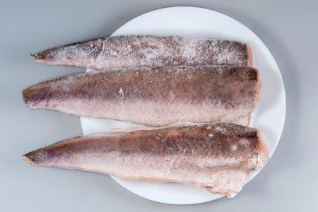 Raw frozen fillets of hake in the ice glaze covered with hoarfrost on plate on a gray background, top viewの写真素材