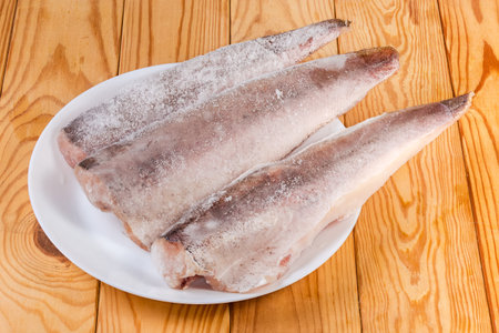 Raw frozen fillets of hake in the ice glaze covered with hoarfrost on white plate on the rustic tableの写真素材