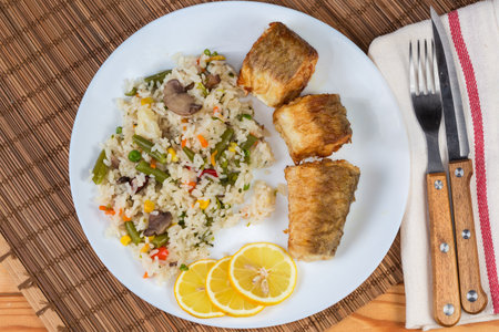 Serving of the pieces of fried Argentine hake, or hake hubbsi with garnish of the boiled mix of rice and vegetables on dish on a bamboo place mat, top viewの写真素材