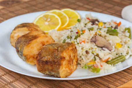 Serving of the pieces of fried Argentine hake, or hake hubbsi with garnish of the boiled mix of rice and vegetables on dish, side view close-up in selective focusの写真素材