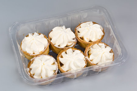 Small tarts, so-called tartlets made with baked shortcrust pastry molds, caramelized pear filling and custard on top in plastic food packaging on a gray backgroundの写真素材