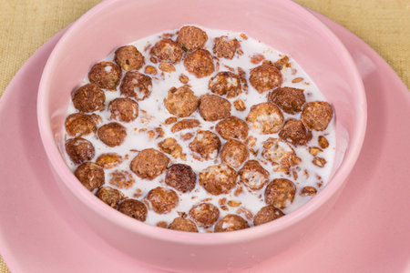 Breakfast cereals in form of mixture of crunchy oat flakes and baked glazed corn balls with cocoa powder drenched in milk in pink bowl, close-upの写真素材