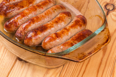 Baked thick pork sausages in natural casing on the old glass casserole pan on a rustic table, fragment close-up in selective focusの写真素材