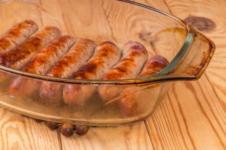Baked thick pork sausages in natural casing on the old glass casserole pan on a rustic table, fragment close-up in selective focusの写真素材
