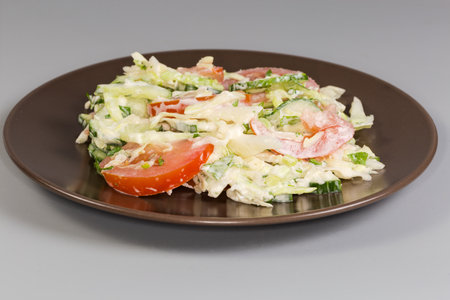 Vegetable salad with sliced fresh tomatoes, cucumbers, onion, young white cabbage with mayonnaise dressing on brown dish on a gray background, side view in selective focusの写真素材