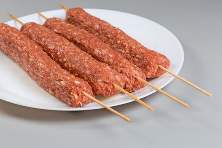 Several raw cooled lula kebab skewered on the wooden bamboo skewers on a white dish on a gray...