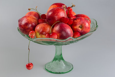 Fresh ripe nectarines and light red sweet cherries on the vintage green glass fruit vase on a leg on a gray background, side viewの写真素材