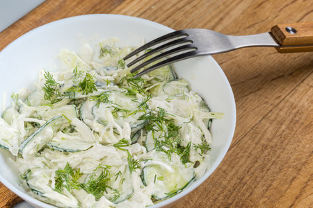 Green vegetable salad made of sliced fresh young cabbage, cucumbers, onion, dill with mayonnaise dressing in the white bowl with fork on a wooden surface close-upの写真素材
