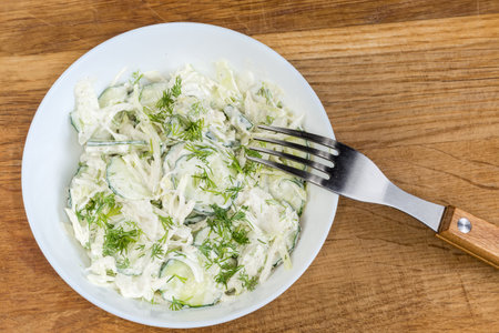 Green vegetable salad made of sliced fresh young cabbage, cucumbers, onion, dill with mayonnaise dressing in the white bowl with fork on an old oak wooden surface, top viewの写真素材