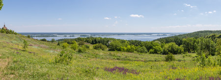 High hilly sloping bank of wide plain river partly partly covered with forest and glade on a foreground in summer sunny day, panoramic viewの写真素材