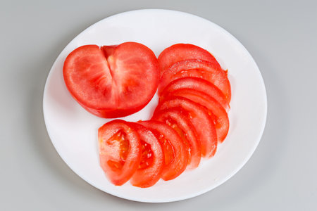 One half and thin slices of the fresh red tomato on dish on a gray backgroundの写真素材