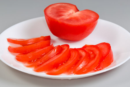 One half and thin slices of the fresh red tomato on dish on a gray background, side view close-up in selective focusの写真素材