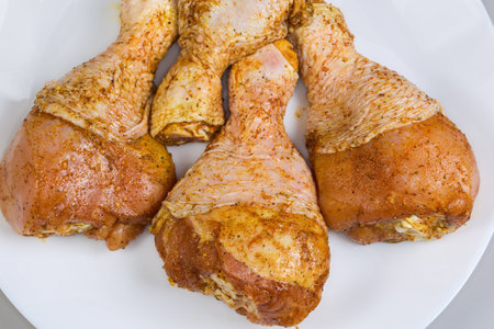 Raw pickled chicken drumsticks sprinkled with different spices on a white dish on a gray background, top view of fragment close-upの写真素材