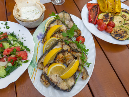 Grilled steaks of the fresh sturgeon with lemon slices and cilantro twigs on the plate, salad of fresh vegetables, baked vegetables on a table, top viewの写真素材