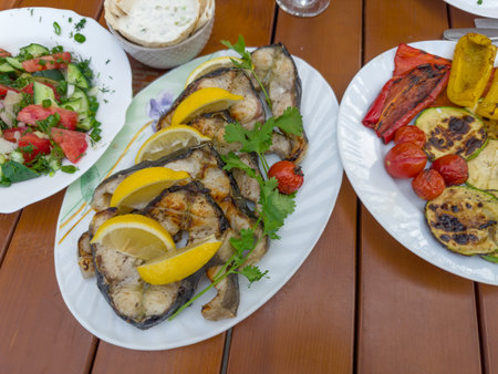 Grilled steaks of the fresh sturgeon with lemon slices and cilantro twigs on the plate, salad of fresh vegetables, baked vegetables on a table, top viewの写真素材