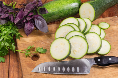 Green zucchini sliced into circles on the wooden cutting board against the same whole zucchini and different greens on the rustic tableの写真素材