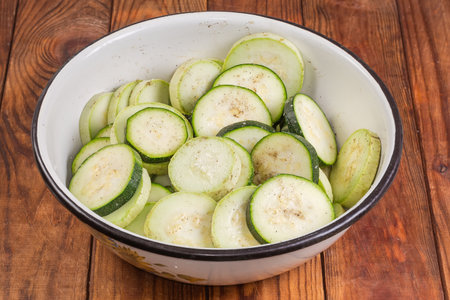 Raw green zucchini and white vegetable marrow sliced into circles and sprinkled with spices in the big kitchen bowl on a rustic tableの写真素材